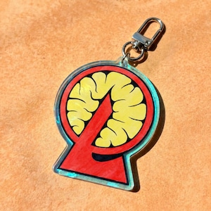 May include: A clear acrylic keychain featuring a circular design with a yellow brain-like graphic centered inside a red triangular and circular frame. The keychain includes a silver-toned metal lobster clasp attached to the top of the acrylic piece.