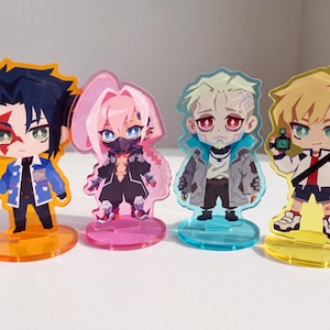 May include: Four acrylic character standees featuring anime-style figures. The first has dark hair and a blue jacket on an orange base. The second has pink hair and a black outfit on a pink base. The third has blonde hair on a blue base. The fourth has blonde hair on a yellow base.