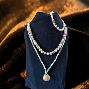 Puede incluir: Juego de collares de varias hebras exhibido sobre un soporte negro. Los collares presentan una mezcla de cuentas blancas, rosas y verdes. El collar inferior tiene un colgante de madera. El fondo es una tela oscura y texturizada.