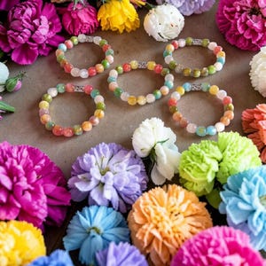 Peut inclure: Plusieurs bracelets colorés à perles rondes, entourés de fleurs vibrantes. Les fleurs sont dans des tons de rose, bleu, jaune et blanc. Les bracelets ont un diamètre d'environ 6,35 cm.