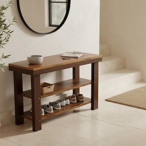 May include: A wooden entryway bench with two shelves, holding several pairs of shoes. The bench has a dark brown frame and a lighter brown top. A small woven basket and a gray bowl sit on the top. A potted plant and a mirror are in the background.