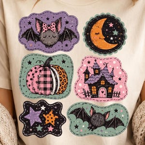 Op de afbeelding: Een crèmekleurig T-shirt met korte mouwen en een grafische print van zes verschillende patches. De ontwerpen bevatten een vleermuis met een roze strik, een halve maan, een pompoen met patronen, een spookhuis, sterren en een vliegende vleermuis.