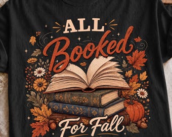 May include: A black short-sleeve t-shirt featuring a graphic print of an open book resting on a stack of two closed books. The design includes autumn leaves, flowers, and a pumpkin, with the text "ALL Booked For Fall" in cream and orange.