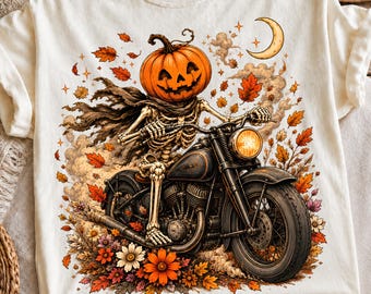 May include: A cream-colored t-shirt featuring a graphic of a skeleton with a jack-o'-lantern head riding a motorcycle. The design includes orange autumn leaves, a crescent moon, and colorful flowers surrounding the motorcycle and skeleton figure.