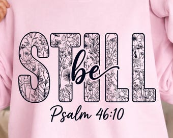 May include: A light pink sweatshirt with the words "STILL be" in a floral design. Below the text is "Psalm 46:10" in a script font. The sweatshirt has long sleeves.