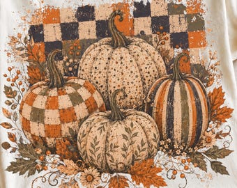 May include: A cream-colored t-shirt featuring a graphic print of four pumpkins with varied patterns including checkered, dotted, and leaf designs. The pumpkins are surrounded by autumn leaves and berries, set against a distressed checkered background.