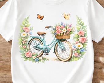 May include: White t-shirt featuring a watercolor illustration of a light blue bicycle with a basket of flowers. The bicycle is surrounded by pink roses, daisies, and butterflies. The shirt has a classic crew neck and short sleeves.