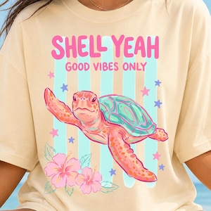 May include: Cream-colored t-shirt featuring a cartoon sea turtle with a teal shell and pink body. The shirt has the text "SHELL YEAH GOOD VIBES ONLY" in pink. Blue and pink stars and flowers decorate the shirt.
