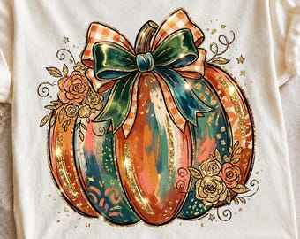 May include: A cream-colored t-shirt featuring a large graphic of a pumpkin with a plaid and solid green bow. The pumpkin is decorated with gold glitter accents, orange and teal brushstroke patterns, and clusters of orange and gold roses.