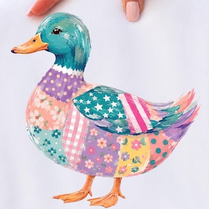 Peut inclure: Un t-shirt blanc avec un motif coloré de canard. Le canard a une tête sarcelle, un bec orange et un corps composé de divers motifs en patchwork, notamment des imprimés floraux, du vichy rose, des étoiles blanches sur fond sarcelle et des pois.