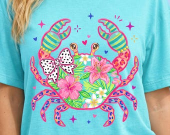 May include: A turquoise t-shirt featuring a colorful crab design. The crab is decorated with pink, green, and yellow floral patterns, a polka-dot bow, and pink flowers. The crab's claws and legs have a vibrant, patterned design. The shirt is a comfortable, casual style.