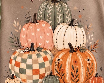 May include: A brown t-shirt featuring a graphic illustration of various pumpkins in shades of green, orange, cream, and peach. The pumpkins display patterns including hearts, checkers, and floral vines, accented by small stars and dots across the design.