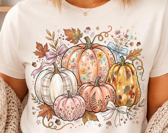 May include: A cream-colored short-sleeve t-shirt featuring a central graphic print of four pumpkins in various colors and patterns, surrounded by autumn leaves, small flowers, acorns, and decorative bows in shades of pink, orange, and blue.