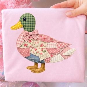 May include: A pink fabric square featuring a patchwork duck design. The duck has a green checkered head, a yellow polka-dot beak, and a pink bow. Its body is made of various patterned fabrics, including floral and gingham prints. The duck has brown feet.
