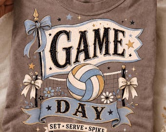 May include: A brown t-shirt featuring a graphic print with the text GAME DAY and SET SERVE SPIKE. The design includes a volleyball, a net, light blue ribbons, bows, and small stars. The shirt has a crew neckline and a standard short-sleeve construction.