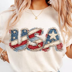 Puede incluir: Camiseta color crema con la palabra "USA" en un diseño patriótico. Las letras están elaboradas con un estilo patchwork, incorporando telas rojas y azules con patrones de estrellas y rayas, y un detalle de botón.
