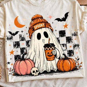 May include: A cream-colored t-shirt featuring a graphic of a ghost wearing an orange beanie with the word SPOOKY, holding a pumpkin-themed coffee cup. The design includes black and white checkered patterns, bats, stars, and two pumpkins.