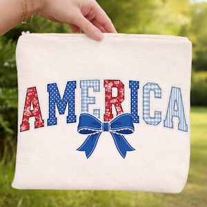Puede incluir: Bolsa con cremallera de color crema con la palabra "AMERICA" en letras estampadas en rojo, blanco y azul. Un lazo azul está centrado debajo de la palabra. La bolsa se sostiene contra un fondo verde borroso.