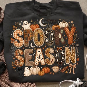 Puede incluir: Una sudadera negra de cuello redondo con el texto SPOOKY SEASON en letras grandes y estampadas. El diseño incluye ilustraciones de fantasmas, calabazas, una luna creciente, telarañas, una taza de café, ramas de canela y lazos en tonos otoñales.