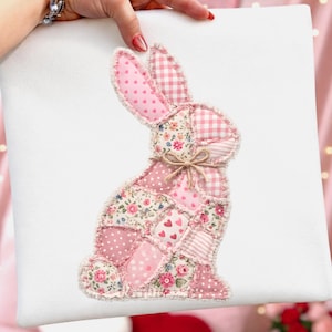 Op de afbeelding: Een wit vierkant kussen met een patchwork konijn ontwerp. Het konijn is samengesteld uit verschillende roze stoffen, waaronder stippen, bloemenprints en ruitpatronen. Een klein strikje is om de nek van het konijn gebonden.