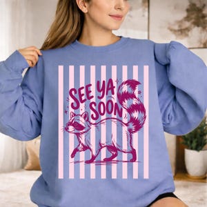 May include: A light blue sweatshirt with a raccoon graphic and the text "See Ya Soon". The raccoon is depicted in shades of gray and pink, with a striped tail. The background features vertical pink stripes.