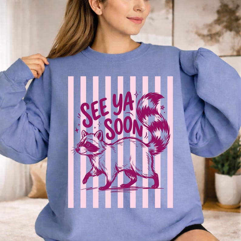 May include: A light blue sweatshirt with a raccoon graphic and the text "See Ya Soon". The raccoon is depicted in shades of gray and pink, with a striped tail. The background features vertical pink stripes.