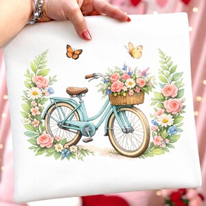 May include: A white cushion cover with a watercolour illustration of a light blue bicycle with a basket of flowers. Pink roses, daisies, and butterflies surround the bicycle, creating a floral design.