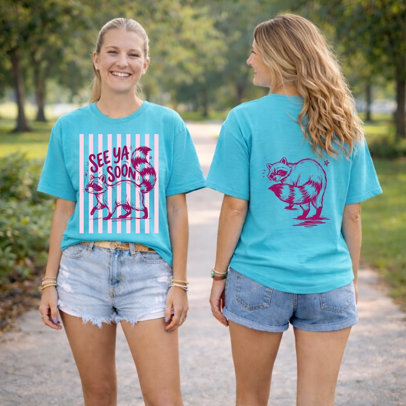 May include: Turquoise t-shirts with a raccoon graphic. The front of the shirt features a raccoon with the text "SEE YA SOON" and vertical pink and white stripes. The back of the shirt has a raccoon graphic.