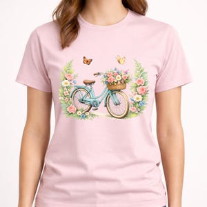 May include: A pale pink t-shirt with a watercolour design of a light blue bicycle with a basket of flowers. The bicycle is surrounded by a floral wreath with pink roses, white daisies, and green leaves. Two butterflies are above the bicycle.