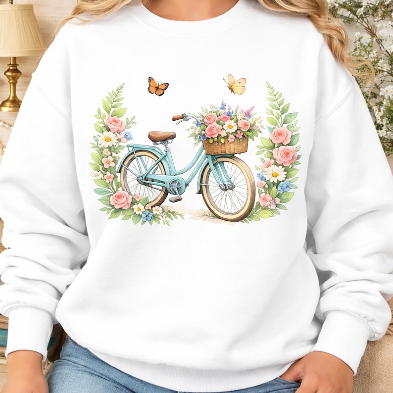 May include: White sweatshirt with a watercolour design of a light blue bicycle with a basket of flowers. The bicycle is surrounded by pink roses, daisies, and green leaves, with two butterflies above.