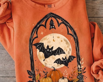 May include: An orange long-sleeve sweatshirt featuring a graphic print of a black gothic archway framing a large full moon. Three black bats are depicted, with two flying and one hanging from the top. Three pumpkins and autumn leaves are at the base.