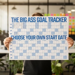Op de afbeelding: Een grote wit en blauwe doeltracker met de tekst "THE BIG ASS GOAL TRACKER" en "CHOOSE YOUR OWN START DATE" in blauw. De tracker wordt voor een persoon in een kantooromgeving omhoog gehouden.