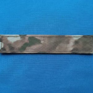 May include: A rectangular fabric strip featuring a camouflage pattern in shades of brown, tan, and dark green. The strip has a woven texture and is finished with stitched edges on both ends, presenting a flat, elongated, and uniform shape.