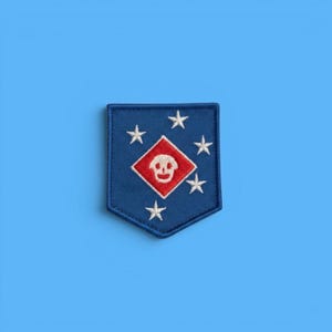 May include: A shield-shaped embroidered patch in dark blue featuring a central red diamond with a white skull icon. The patch is decorated with five white embroidered stars arranged around the central diamond shape on the blue fabric background.