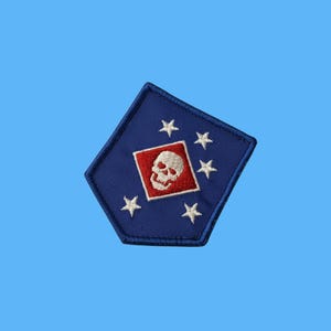 May include: A pentagonal embroidered patch featuring a blue background with a central red diamond containing a white skull. Five white stars are arranged in a curved pattern around the diamond, and the entire patch has a dark blue stitched border.