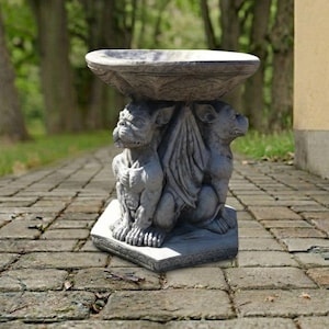 May include: A gray stone birdbath featuring a wide, circular basin supported by a pedestal base. The base is sculpted with two detailed gargoyle figures, each with wings tucked against their bodies, standing on a hexagonal stone platform.