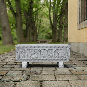 May include: A rectangular light gray stone planter featuring a relief carving of cherubs in various poses across the front panel. The planter is framed by a braided border design and is supported by two short, carved feet on a paved surface.