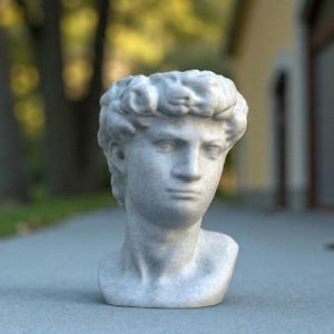 May include: A small, detailed bust of a classical male figure, likely a sculpture. The bust is a light gray color, with visible texture and shadows that highlight the facial features and curly hair. The sculpture is set against a blurred outdoor background.