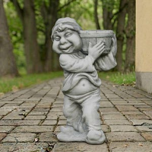 May include: A gray stone garden statue of a gnome figure wearing a cap and loose clothing, holding a small bucket with both hands. The figure has a smiling expression, a rounded belly, and is standing on a solid base, crafted with a textured finish.