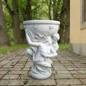 May include: A light gray stone planter featuring a sculpted base of two figures embracing and holding up the bowl. The bowl has a smooth rim decorated with small, evenly spaced circular floral carvings around the exterior circumference.
