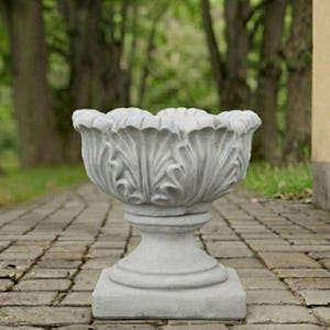 May include: A light gray stone planter featuring a wide, bowl-shaped top with a scalloped, leaf-like rim design. The planter rests on a circular pedestal base that connects to a square foundation, showcasing a textured, weathered finish throughout.
