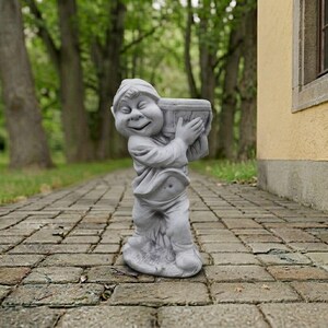 May include: A gray stone garden statue of a smiling figure wearing a cap and loose clothing, holding a cylindrical bucket or container above its head with both hands. The figure has a rounded belly and stands on a textured, uneven stone base.