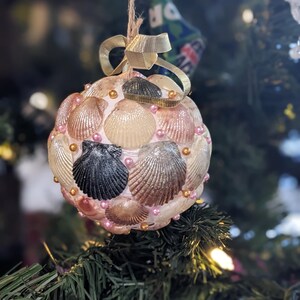 May include: A handcrafted Christmas ornament, spherical in shape, adorned with various seashells in shades of white, tan, and brown. The ornament is accented with pink beads and a gold ribbon, suspended by twine, creating a coastal holiday theme.