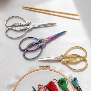 Könnte beinhalten: Drei dekorative Stickscheren in Silber, schillernd und Gold, mit verzierten Griffen. Ein Stickrahmen mit floraler Stickerei und Garnrollen sind ebenfalls zu sehen.