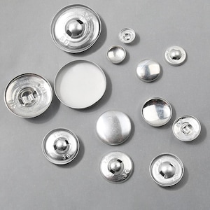 May include: An assortment of silver metal snap fasteners in various sizes. The snaps have a shiny, reflective surface and are arranged on a gray background. Some snaps have the number "36" stamped on them.