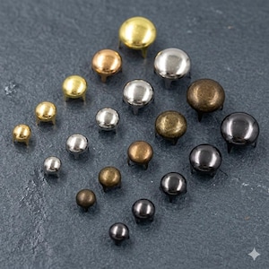 May include: A collection of metal dome rivets in different sizes and finishes, such as gold, silver, bronze, and black. These decorative rivets have a rounded head and three prongs for attachment, suitable for leathercraft and DIY projects.