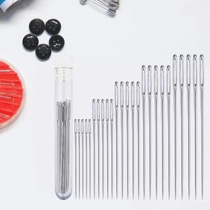 May include: A collection of sewing supplies, including a clear tube filled with needles, a set of black buttons, and a variety of sewing needles. The needles are of varying lengths, and the buttons have four holes. The items are arranged on a white surface.