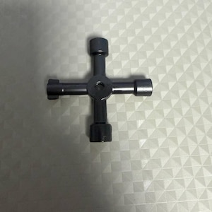 May include: A dark gray, cross-shaped lug wrench. The tool has four arms with different sized sockets, and a central circular hub. The wrench is made of metal and is designed for automotive use.