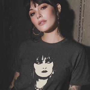 May include: A charcoal gray short-sleeved t-shirt featuring a high-contrast, cream-colored graphic print of a person with dark, voluminous hair, heavy eyeliner, and a beaded necklace. The shirt has a crew neckline and a standard, straight-cut fit.