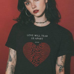 Puede incluir: Una camiseta negra de manga corta con el texto blanco LOVE WILL TEAR US APART AGAIN sobre un gráfico rojo en forma de corazón formado por líneas horizontales que recuerdan a un patrón de ondas sonoras. La camiseta tiene cuello redondo.